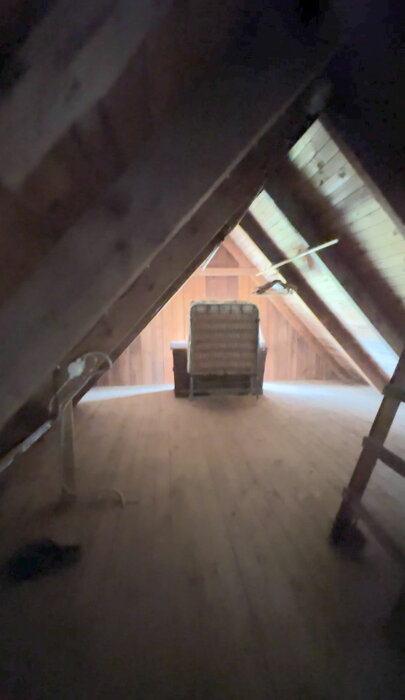 Vindsutrymme med trätak och takstolar mot en kort vägg. En stol är placerad intill en låda vid väggen.