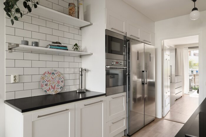 Kök med vit inredning, inbyggd ugn och mikrovågsugn bredvid kyl och frys, hyllor med dekorativa föremål och kaklad vägg.