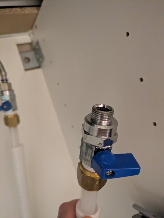 Vattenrörsanslutning med blå ventil, metallkoppling och vit pexslang, monterad i ett skåp.