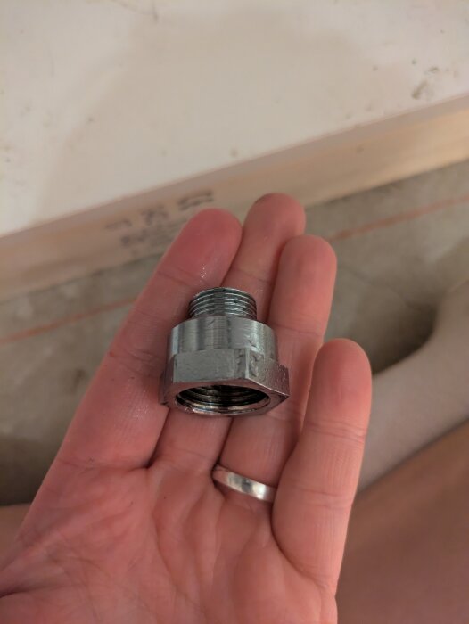 En hand håller en metallkoppling eller adapter med gängor, troligen en del av ett VVS-system, mot en bakgrund av golv och trä.