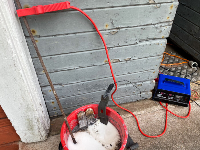 Elektrolyskärl med röda kablar anslutna till ett batteriladdare och en stång i en hink med lösning, mot en vägg.