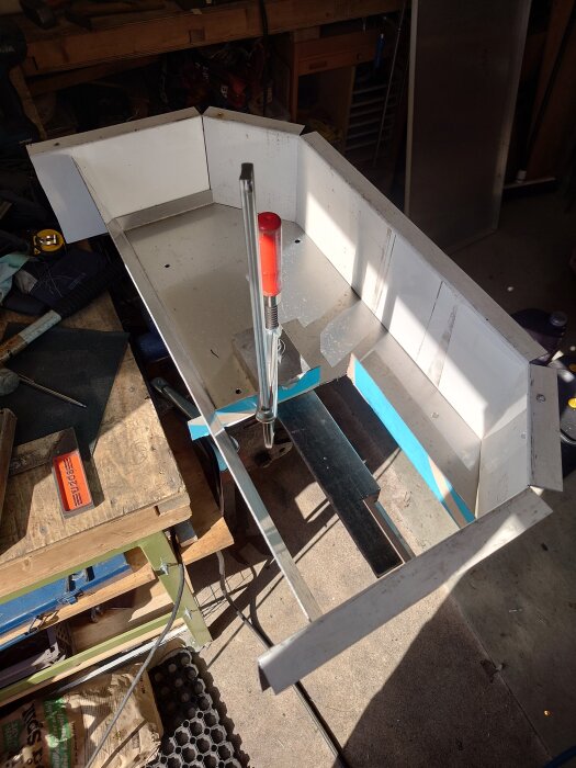 Arbetsbänk med upphöjt aluminiumföremål i garage, omgivet av verktyg och arbetsmaterial.