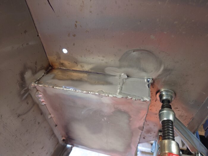 Svetsad aluminiumplåt med borrhål och en fastsatt klämma.