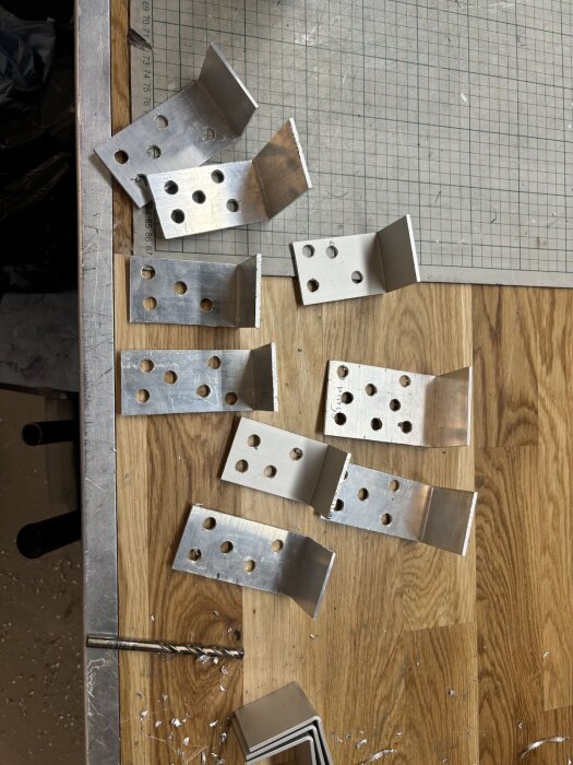 Vinkelfästen i aluminium med borrade hål, placerade på ett träbord. Verktyg och metallspån syns också på bordet.
