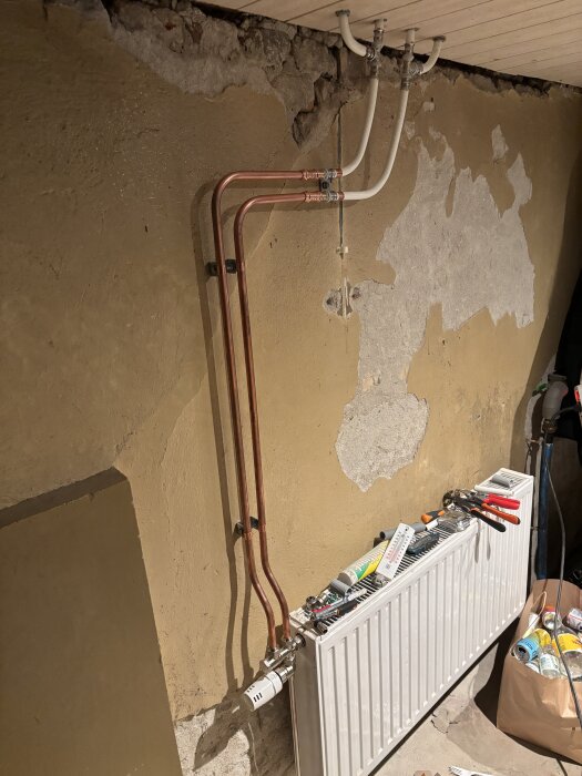 Rördragning av koppar och vita rör i tak och vägg ovanför ett element med verktyg och material, omgiven av sliten vägg med flagnande färg.