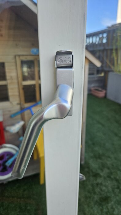 Handtag på altandörr i silver, monterat på vit dörrkarm, med inskription "K-14 K-26/K-34", del av en trasig dörrbroms.