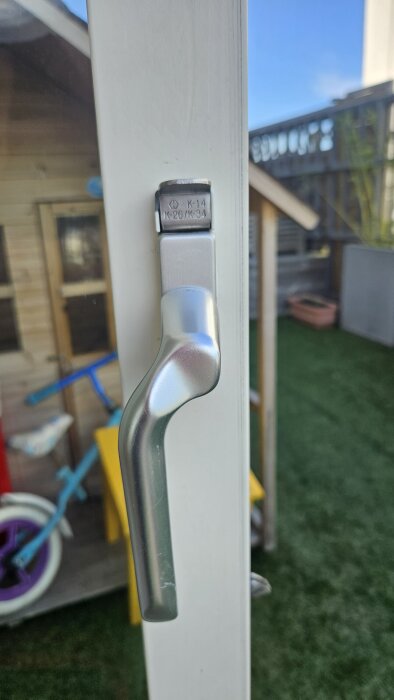 Silvrigt handtag på altandörr med glapp och funktionsproblem, mot bakgrund av en lekstuga och cykel utomhus.