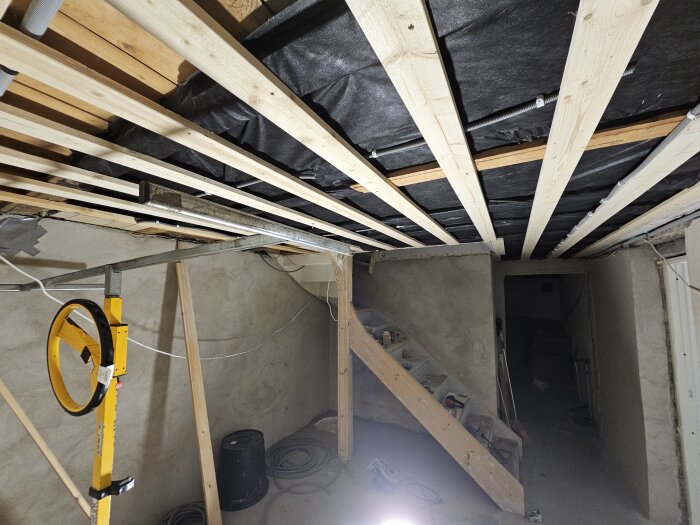 Renovering av källartak med exponerade träreglar och isolering samt en gul bygglampa i förgrunden. En trappa leder till ett mörkt utrymme.