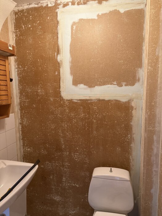 Vägg med bortskrapade limrester i ett badrum, synliga fläckar och materialförlust. Toalett och handfat i förgrunden.