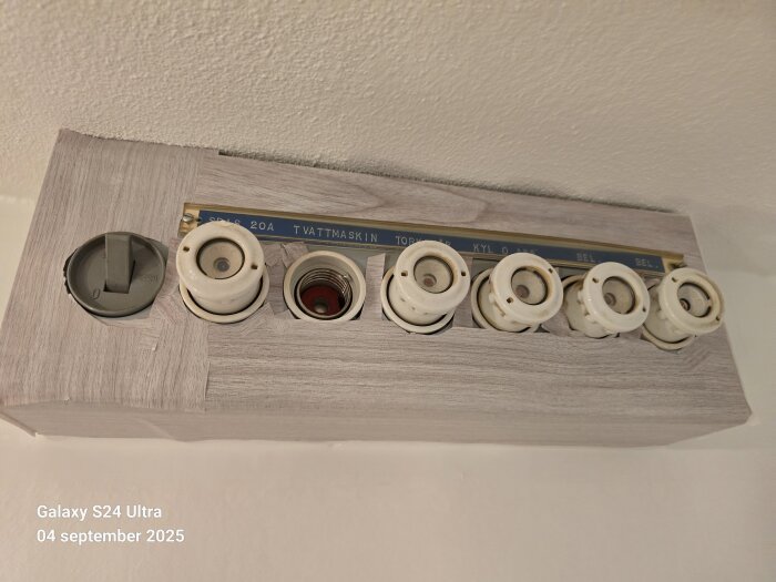 Gammalt proppskåp i badrum med flera porslinssäkringar omgivna av träramar, varav en märkning lyder "TVÄTTMASKIN".