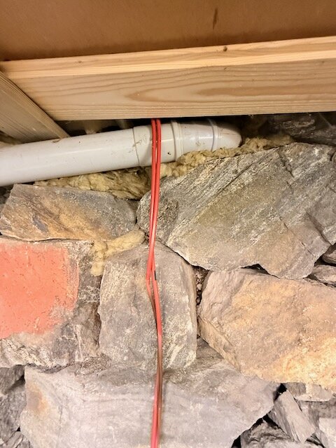 Röd värmekabel och vit rörledning i stenmiljö under en träbalk i en källare.