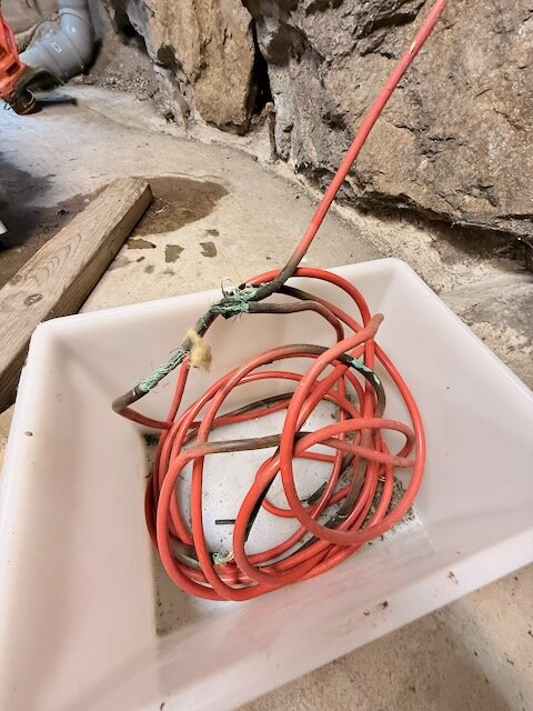 Röd värmekabel i källare, delvis oisolerad, ligger i en vit plastbalja. Bakgrund av betonggolv och stenväggar syns.