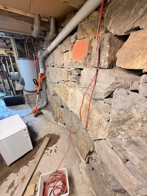 Röd värmekabel längs stenvägg i sommarhusets källare, delvis isolerad och ledande till vattenledningar och varmvattenberedare.