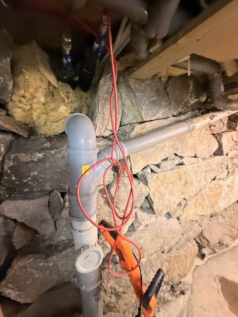 Röd värmekabel slingrande runt gråa rör i en källare med stenväggar, isolering synlig ovanför.