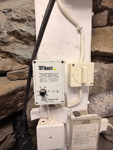 En Termonic-regulator monterad på en träskiva i en källare, med synliga kablar och stenvägg i bakgrunden.