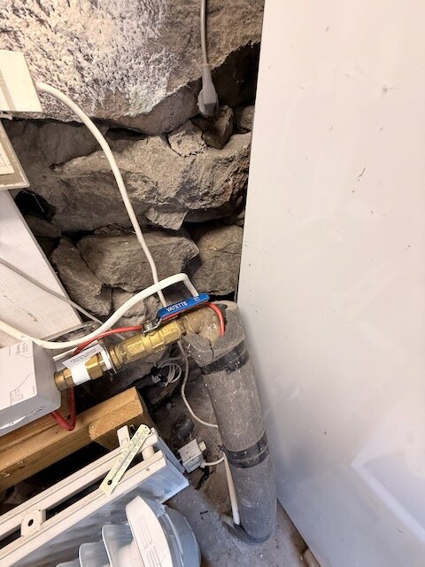 Röd värmekabel ovanpå vattenrör, delvis isolerad och oisolerad, omgiven av stenar och installationsmaterial i en källare.