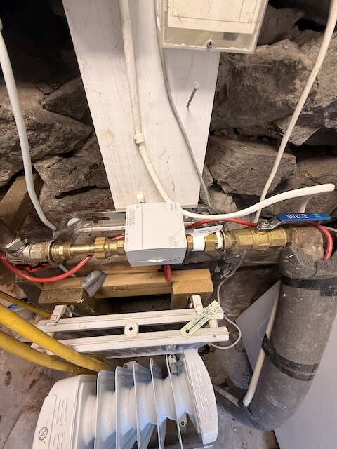 Röd värmekabel och rör i en källare, kabel delvis bränd och kopplad längs rör, termostat synlig, stenar och olika rörmaterial i bakgrunden.