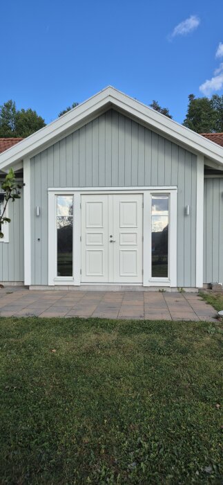 Fasaden av ett enplanshus med dubbeldörr och runtomstående fönster, med grönt gräs och träd i bakgrunden under en blå himmel.