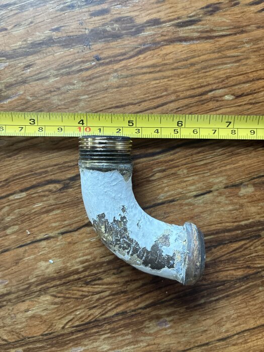 En gammal VVS-rörböj med gängor, bredvid ett måttband på ett träbord.