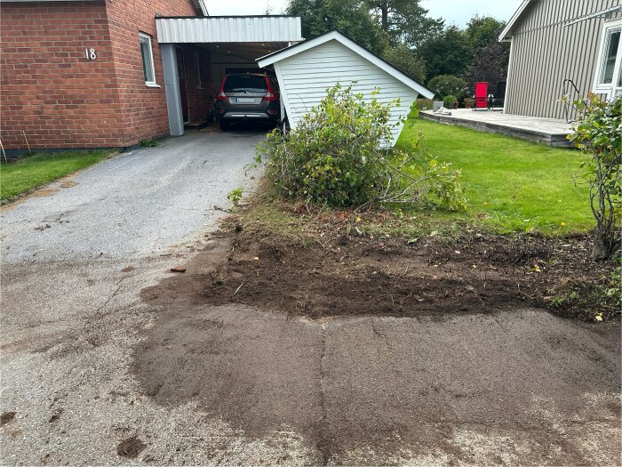 Del av en uppfart med borttagen jord, bredvid grönska och en parkerad bil under carport.