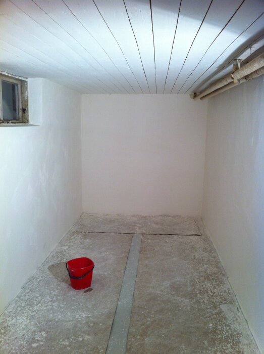 Renoverad källare med synlig radiatorslinga i taket och en röd hink på betonggolvet. Väggar och tak är vita.