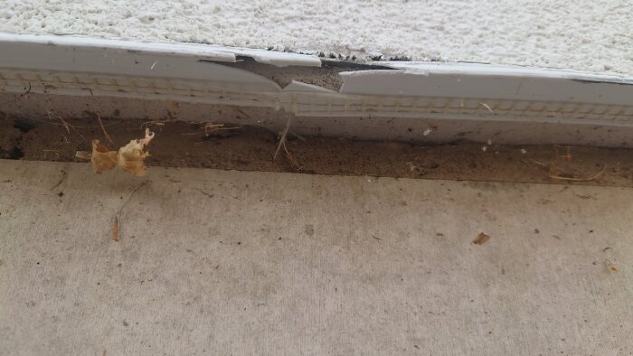 Skadad avvattningslist med ca 15 cm spricka på en byggnadsyta, omgiven av smuts och skräp.