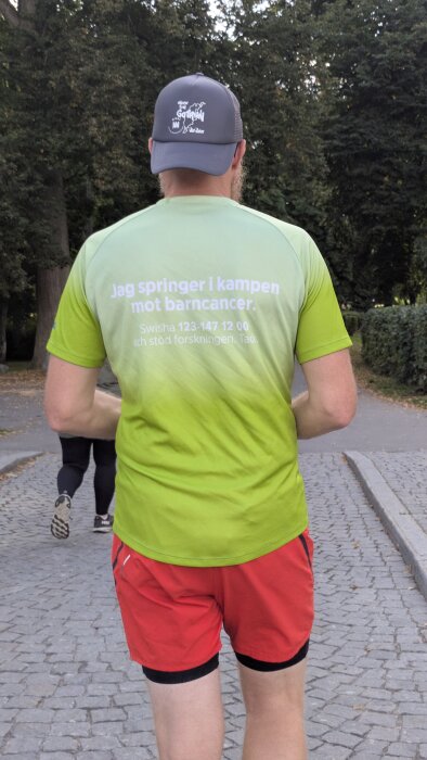 Man i grön tröja och röd shorts springer på en grusväg. Texten på tröjan stödjer kampen mot barncancer.