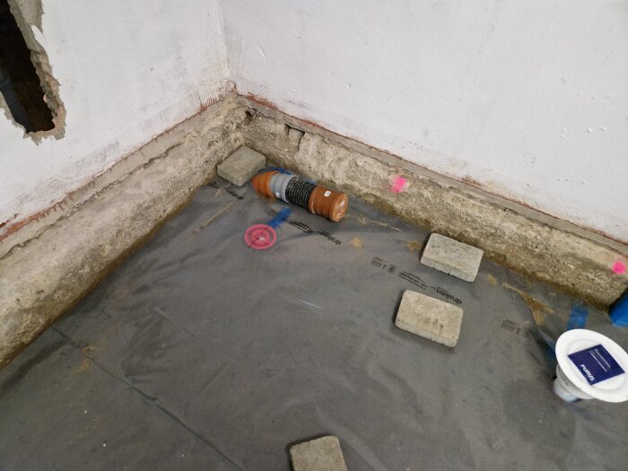 Betonggolv i källare under renovering med synligt rör som förberedelse för radonsug, omgiven av några tegelstenar och plastöverdrag på golvet.
