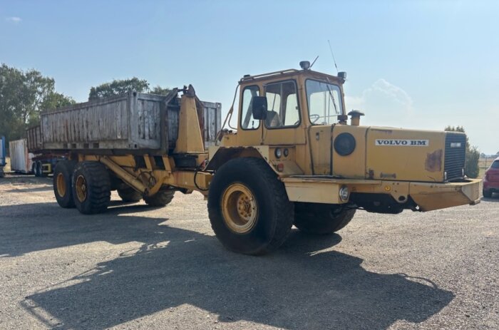 Gul Volvo BM traktorladdare med släp står parkerad på en grusplan under en klar himmel.