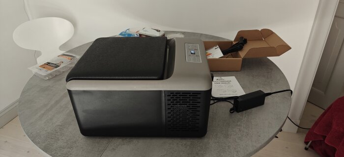 Portabel kyl med ett lock på ett runt bord, omgiven av förpackningar och en manual.