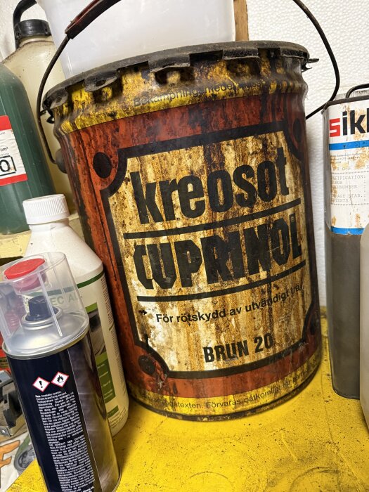 Rostig hink med texten "kreosot CupRinol", avsett för träskydd, omgiven av andra kemikaliebehållare på hylla.