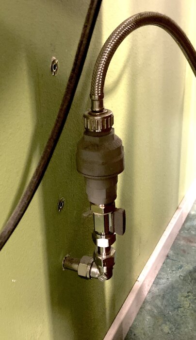 VVS-komponent monterad på en grön vägg, med flexibel slang och kopplingar, okänt ändamål.