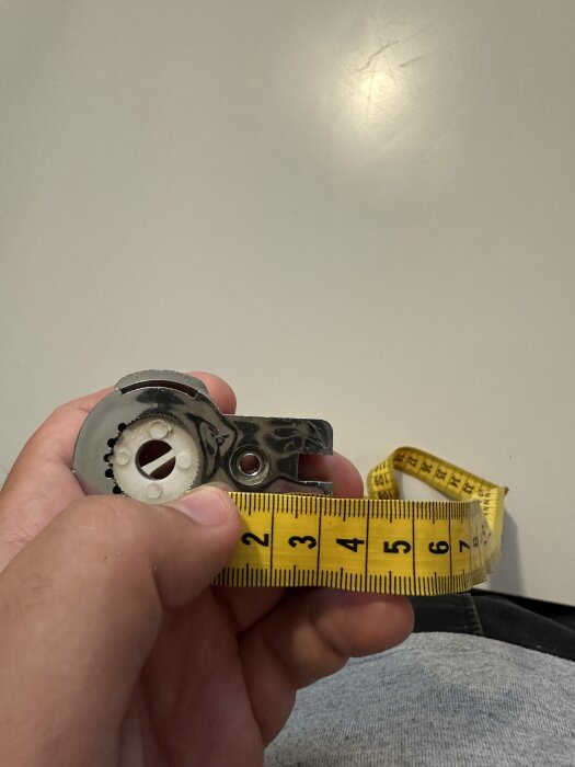 En hand håller i en metalldel kopplad till duschdörrar av märket Elsa, intill ett måttband som visar cirka 7 cm.