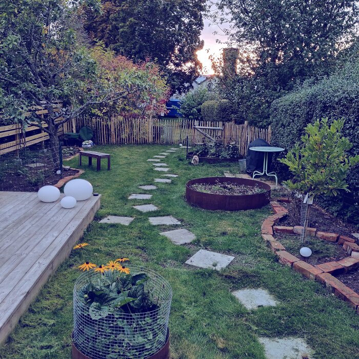 Liten trädgård med stenlagda gångar och planteringar, omgiven av staket och buskar, med en liten uteplats till vänster och en rund eldskål i mitten.