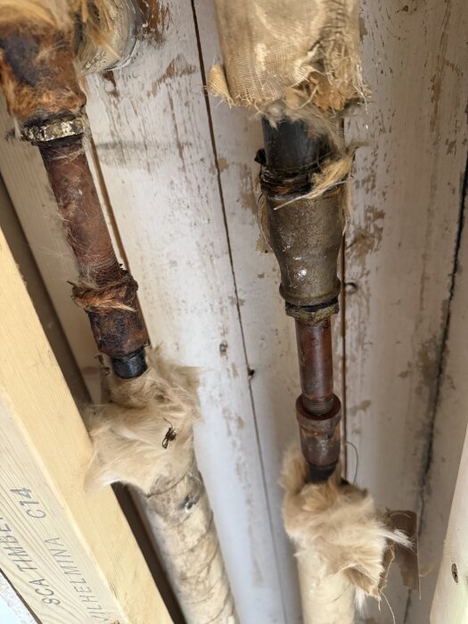 Rostiga vattenrör med isolering mot en gammal trävägg.