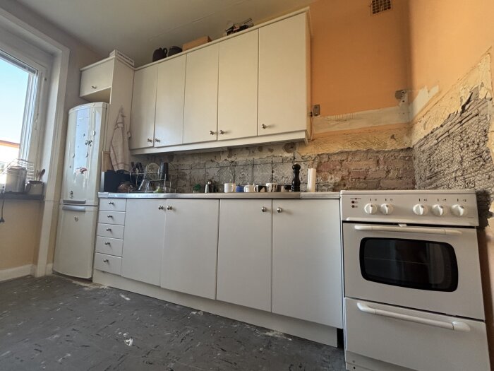 Kök under renovering med borttaget kakel, exponerade tegelväggar och vita skåp, samt en vit spis på höger sida.