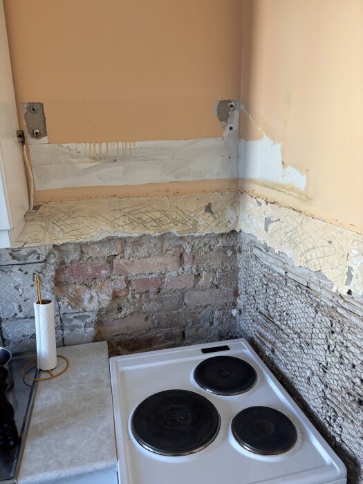 Kök med borttaget kakel, delvis nedbruten vägg, synliga tegelstenar och en spis. Bänkskiva intill spis med hushållspapper på.