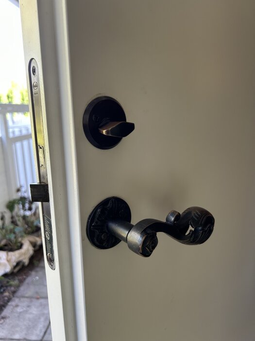 Ytterdörr med gammal Boda 444-låskista och dekorerat dörrhandtag i metall, fotograferad från insidan med utsikt mot trädgård.