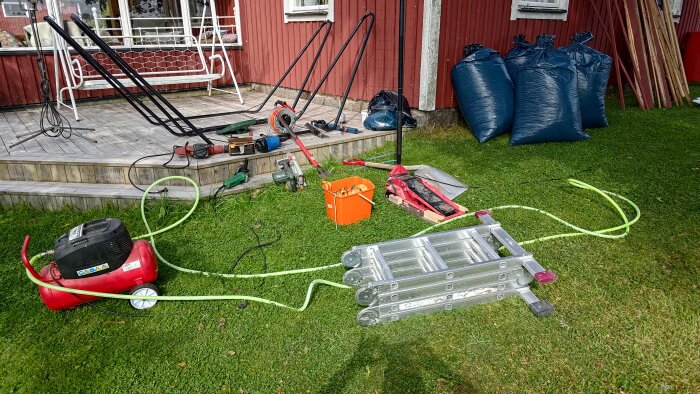 Verktyg och utrustning spridda på gräsmatta framför en altan, inkluderande kompressor, stege och diverse byggredskap.