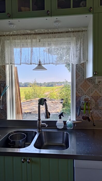 Utsikt från köksfönster med lantlig vy, synlig diskho och kran i förgrunden, samt ett borttaget förråd därute.