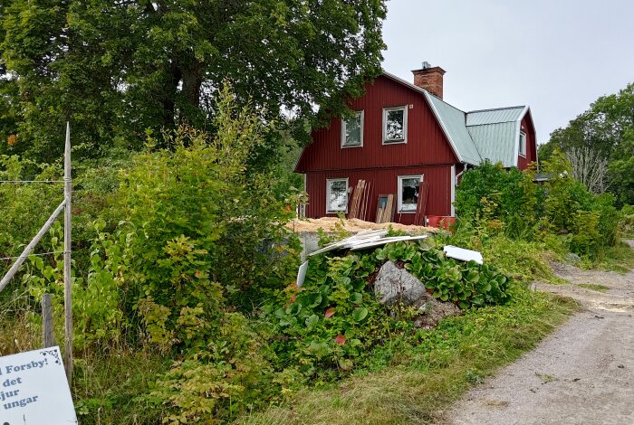 Rött hus på landsbygden med renoveringsmaterial vid sidan om, omgiven av grönska och grusväg.