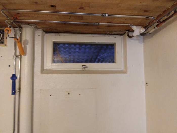 Fönster i källartak med rördragning ovanför, vattenrör och avloppsrör, träpanel i taket och en vitmålad vägg.