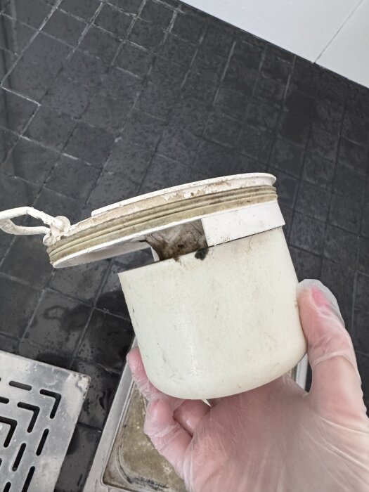 En hand med plasthandske håller i ett vattenlås, delvis öppet, med synligt smuts. Golvbrunn och kaklat badrumsgolv i bakgrunden.