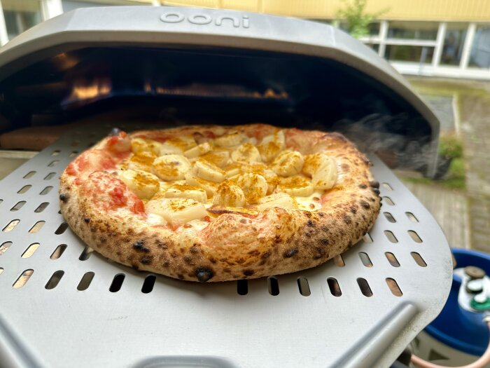 En pizza med ost och potatis gräddas i en portabel pizzaugn av märket Ooni, utomhusscen med träaltan i bakgrunden.