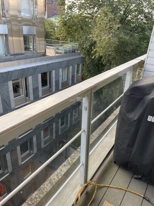 Balkong med glasräcke där vattendroppar från ovan riskerar att spridas, med vy över byggnader och träd i bakgrunden.
