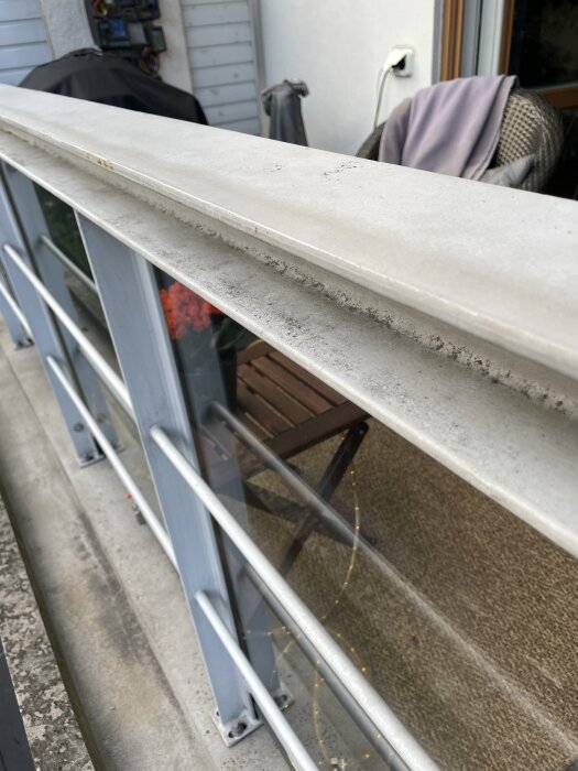 Balkongräcke med överliggare där vatten samlas och sprids, visas med utemöbler i bakgrunden och en betongplatta nedanför.