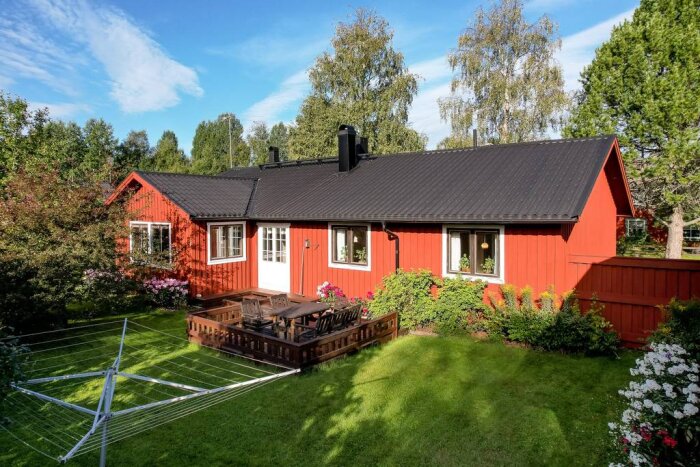 Rött enplanshus med svart tak, flankerat av träd och buskar. Trädäck med utemöbler och tvättställning på gräsmattan i förgrunden.