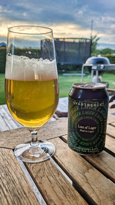 Ett glas lageröl bredvid en svart burk med etiketten "Lots of Lager Modern Lager" på ett träbord utomhus.