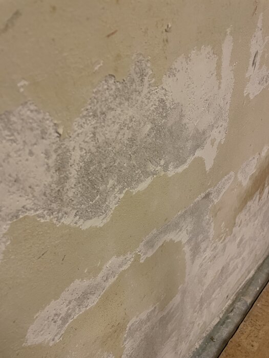 Yttervägg i källare med fuktskador och bortskrapat putslager, visar flera ljusa och mörka fläckar och några större hål som behöver lagas.