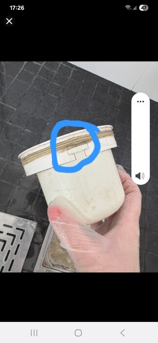 En hand i plasthandske håller en vit behållare med lock, markerad med blå ring runt en flärp som fäster locket.